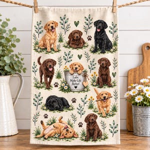 May include: A cream-colored kitchen towel featuring illustrations of yellow, chocolate, and black Labrador puppies in various poses. The center shows a bucket with the text "Labs Make Life Better" surrounded by daisies, green leaves, and paw prints.