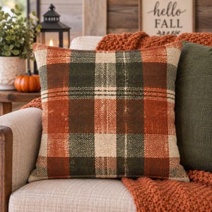 Könnte beinhalten: Ein quadratisches Zierkissen mit einem Karomuster in den Farben gebranntes Orange, Waldgrün und Creme. Der Stoff hat eine texturierte, gewebte Optik mit einem abgenutzten, verwitterten Aussehen entlang der Linien des Schottenmusters.