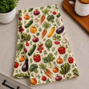 May include: A rectangular kitchen towel featuring a repeating pattern of various colorful vegetables including carrots, broccoli, bell peppers, onions, eggplants, zucchini, radishes, and artichokes printed on a light cream-colored fabric background.
