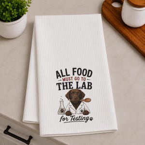 May include: A white waffle-weave kitchen towel featuring a graphic of a brown Labrador dog wearing a lab coat, holding a wooden spoon, and standing next to a beaker. The text reads: ALL FOOD MUST GO TO THE LAB for Testing with paw prints.