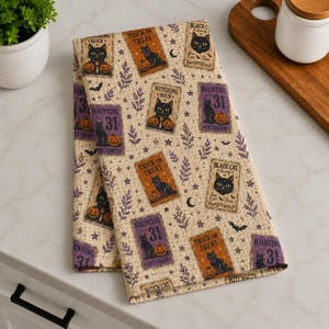 May include: A beige waffle-weave kitchen towel featuring a repeating pattern of rectangular labels with black cats, pumpkins, and stars. The text includes "Witching Hour," "Trick or Treat," "Haunting 31 October," and "Black Cat Society" in various fonts.