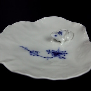 May include: A white ceramic dish with a scalloped edge, featuring a small frog figurine and blue floral detailing. The dish is set against a black background, highlighting its delicate design.