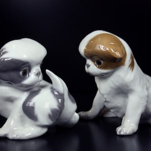 以下が含まれることがあります： 2つのセラミック犬の置物。1つは白とグレーの模様、もう1つは白と茶色の模様です。どちらも座った姿勢で、頭を傾けています。置物は光沢のある仕上げで、黒い背景に配置されています。