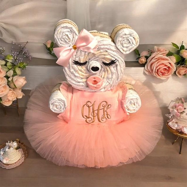 May include: A diaper cake shaped like a bear with a light pink tutu, a matching bow on its ear, and a pacifier. The bear wears a peach-colored top with the embroidered monogram QHK in brown thread. The bear has closed eyes with long lashes.