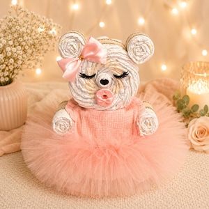 May include: A diaper cake shaped like a bear, constructed from rolled white diapers. The bear features a pink satin bow on its ear, long black eyelashes, and a pink pacifier. It wears a soft pink muslin dress with a layered tulle tutu skirt.