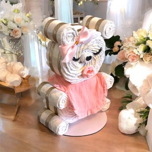 May include: A diaper cake shaped like a bear, constructed from rolled white diapers with gold glitter ribbon accents. The bear features a pink satin bow, a pink pacifier, and a soft pink fabric bib. It has closed eyes with long black eyelashes.