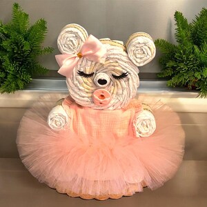 May include: A bear-shaped diaper cake constructed from rolled white diapers, featuring a light pink tulle tutu, a matching pink bow on the head, and a pink pacifier. The bear has closed eyes with long black eyelashes and round ears.