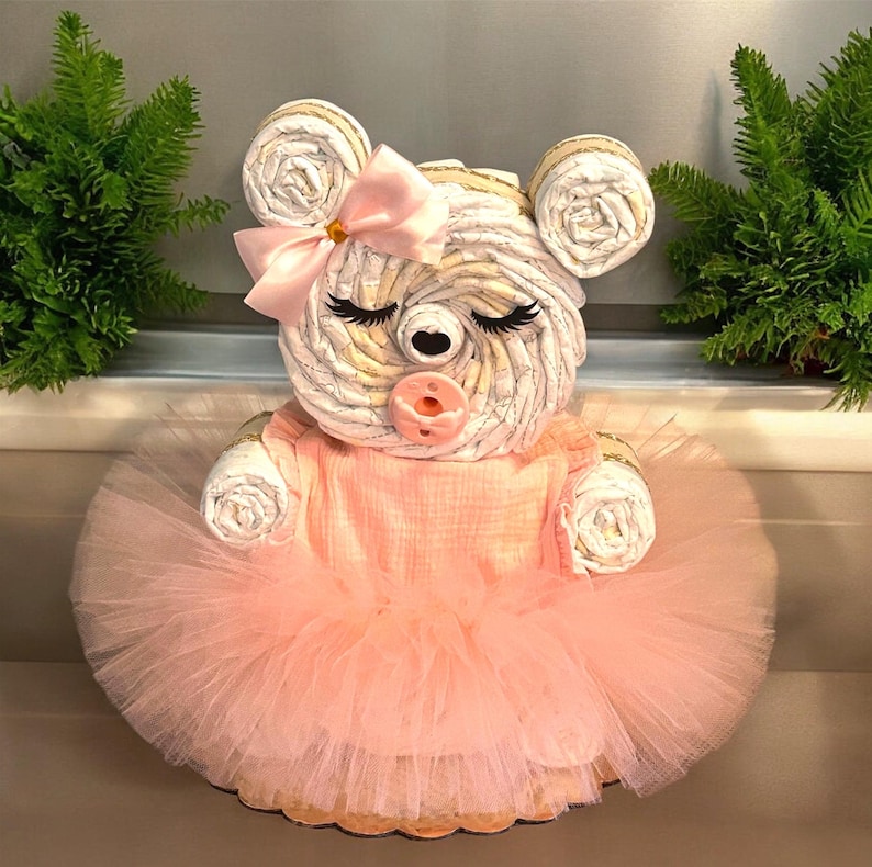 May include: A bear-shaped diaper cake constructed from rolled white diapers, featuring a light pink tulle tutu, a matching pink bow on the head, and a pink pacifier. The bear has closed eyes with long black eyelashes and round ears.