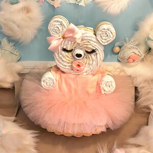 May include: A diaper cake shaped like a bear, constructed from rolled white diapers. The bear features a pink satin bow on its ear, a pink pacifier, and closed eyes with long lashes. It wears a peach-colored fabric top and a layered tulle skirt.