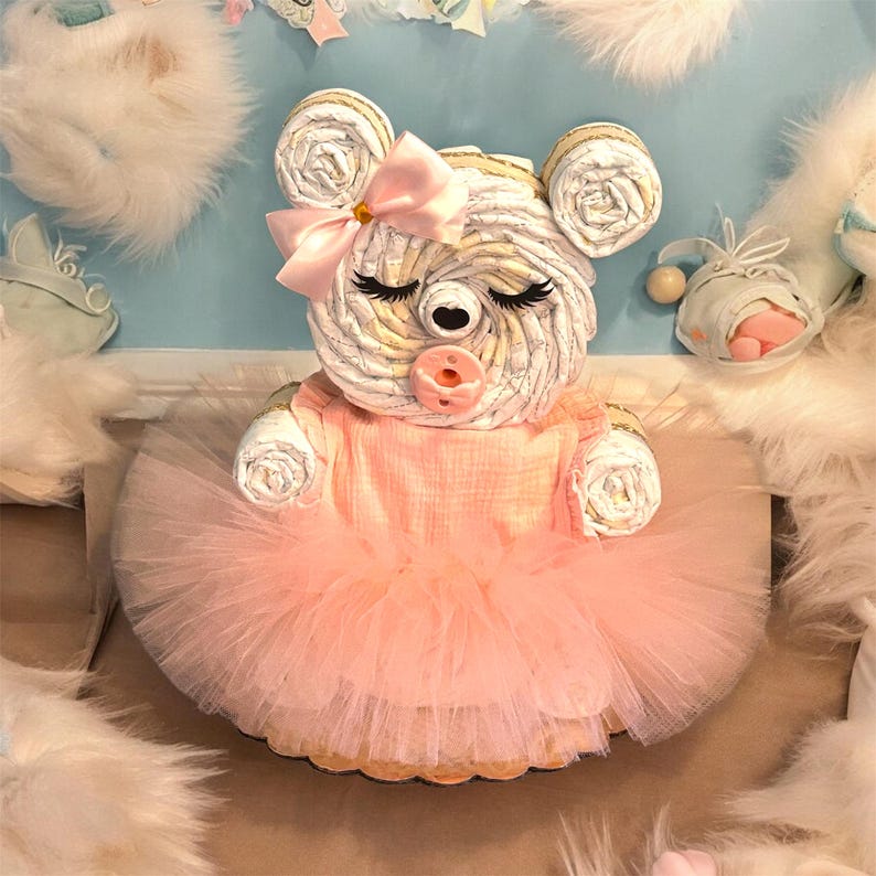 May include: A diaper cake shaped like a bear, constructed from rolled white diapers. The bear features a pink satin bow on its ear, a pink pacifier, and closed eyes with long lashes. It wears a peach-colored fabric top and a layered tulle skirt.