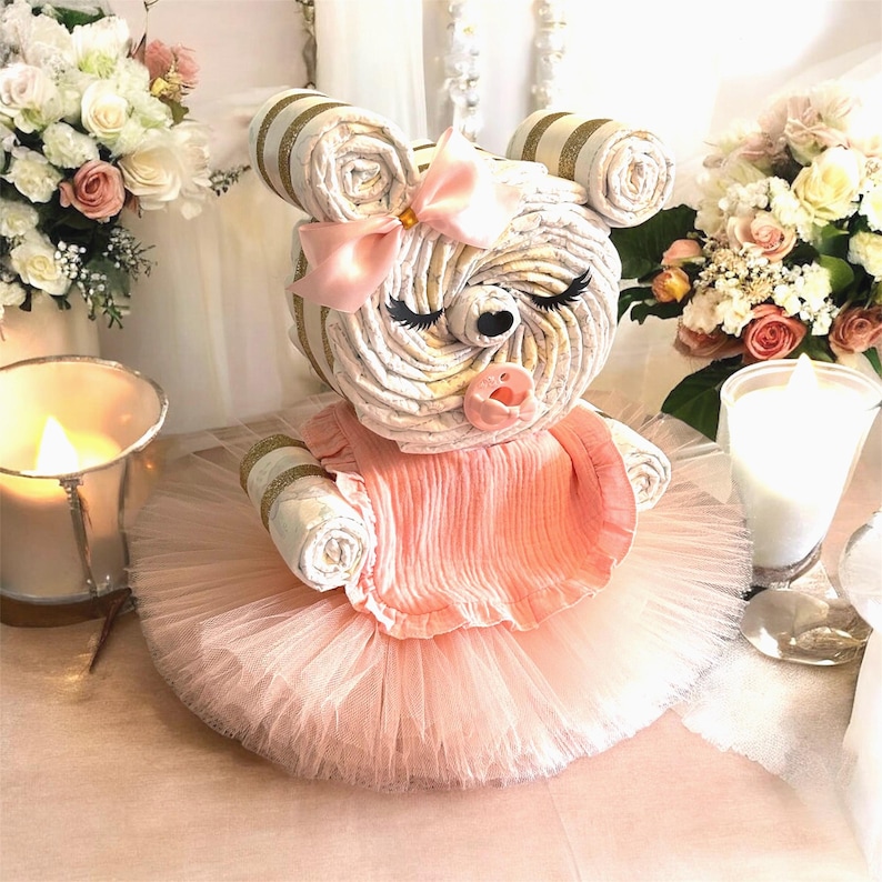 May include: A diaper cake shaped like a bear, constructed from rolled white diapers with gold accents. The bear features a pink satin bow, a pink pacifier, and closed eyelashes. It wears a textured peach-colored bib and a layered peach tulle skirt.