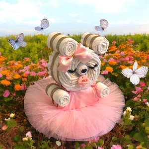 May include: A diaper cake shaped like a bear, featuring rolled white diapers with gold glitter bands, a pink satin bow, a pink pacifier, and a pink tulle tutu skirt. The bear has closed eyes with long black eyelashes and a small black nose.