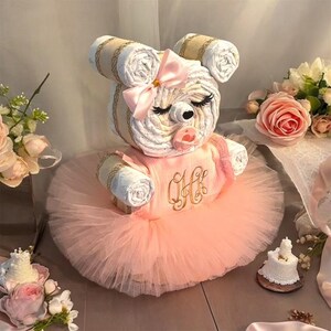 May include: A diaper cake shaped like a bear with a pink tulle skirt, a pink satin bow on its head, and a pacifier. The bear has closed eyes with long lashes and a gold embroidered monogram on its chest. The ears are made of rolled white diapers.