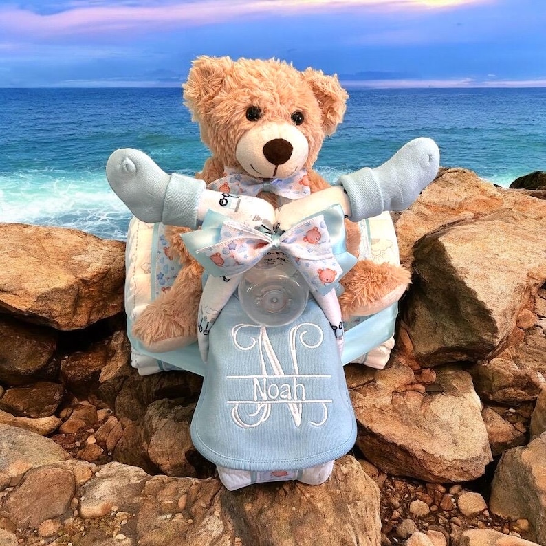 K&ouml;nnte beinhalten: Eine blaue und wei&szlig;e Windeltorte mit einem Teddyb&auml;r, der auf einem Roller f&auml;hrt. Die Torte ist mit einer Schleife verziert und der Name "Noah" ist auf der Vorderseite gestickt.