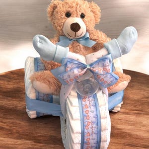 K&ouml;nnte beinhalten: Eine Windeltorte in Form eines blauen Dreirads mit einem braunen Teddyb&auml;ren, der oben sitzt. Das Dreirad ist mit blauem und wei&szlig;em Band und einer blauen Schleife verziert. Der Teddyb&auml;r tr&auml;gt blaue Socken.