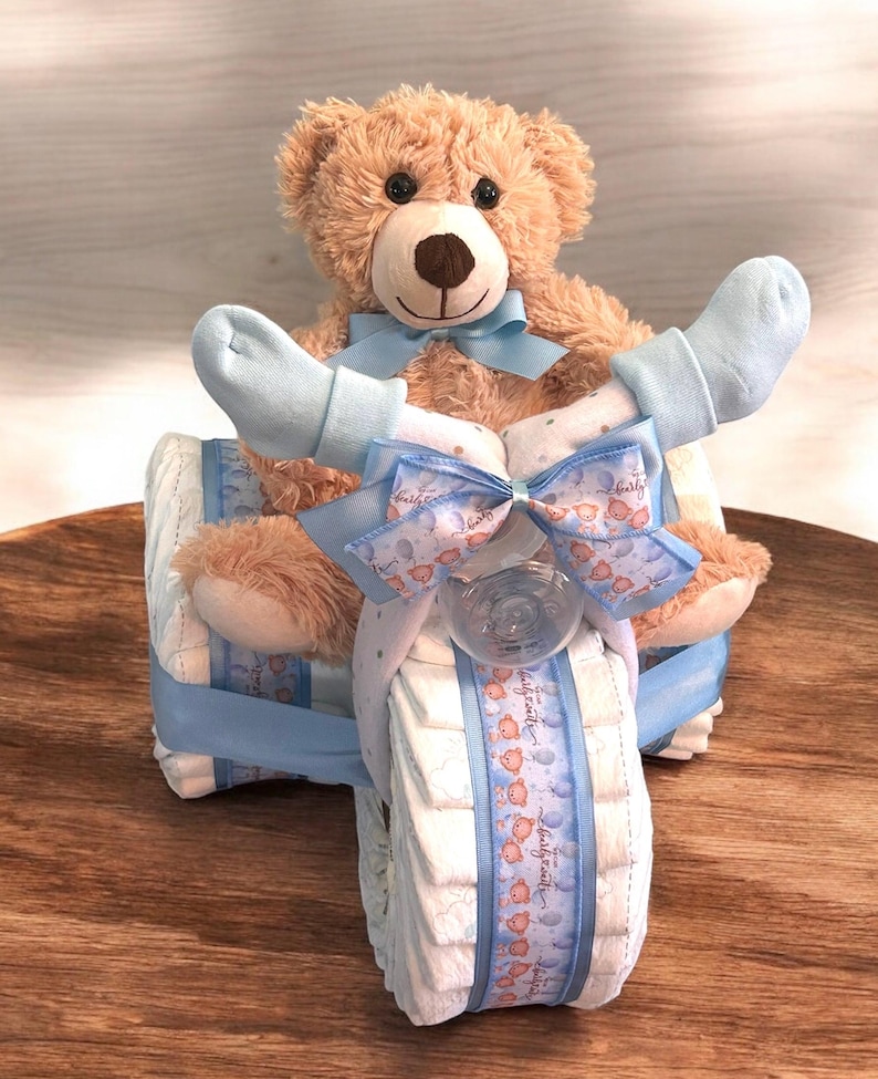 K&ouml;nnte beinhalten: Eine Windeltorte in Form eines blauen Dreirads mit einem braunen Teddyb&auml;ren, der oben sitzt. Das Dreirad ist mit blauem und wei&szlig;em Band und einer blauen Schleife verziert. Der Teddyb&auml;r tr&auml;gt blaue Socken.