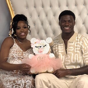 May include: A diaper cake shaped like a teddy bear, featuring white rolled diapers, a pink tulle tutu, a pink pacifier, and a pink bow. The tutu is embroidered with the gold script text 'Oh Baby'. The bear has closed eyes with long lashes.