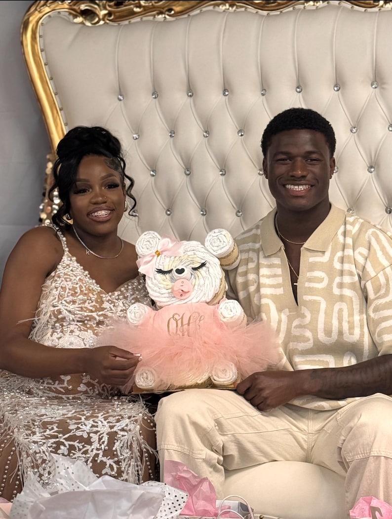 May include: A diaper cake shaped like a teddy bear, featuring white rolled diapers, a pink tulle tutu, a pink pacifier, and a pink bow. The tutu is embroidered with the gold script text 'Oh Baby'. The bear has closed eyes with long lashes.