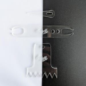 May include: Clear acrylic craft tools arranged against a black and white background. The tools include a needle threader, a loop tool, and a tool with a notched edge. The tools have the text "Handyly" on them.
