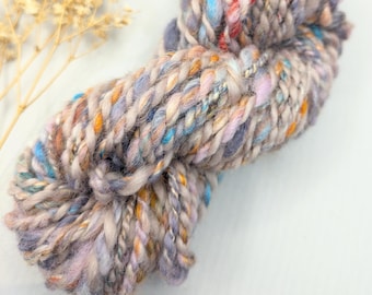 May include: A skein of hand-dyed yarn in shades of purple, blue, orange, and pink. The yarn is thick and fluffy, with a textured appearance.