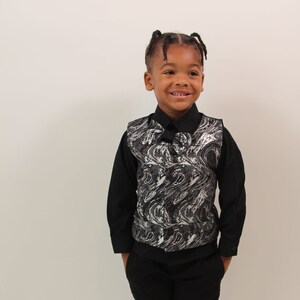 May include: A young boy wearing a black shirt and black pants with a silver and black patterned vest. He is smiling and looking to the right.