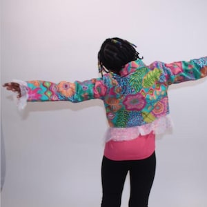 May include: A colorful, patterned jacket with floral and leaf designs in shades of pink, blue, green, and yellow. The jacket has long sleeves with white lace cuffs and a ruffled hem. The person is wearing a pink shirt and black pants.
