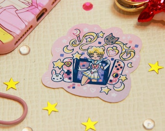 Puede incluir: Una pegatina rosa y blanca con un personaje de dibujos animados con un traje de marinero sosteniendo un mando de videojuegos. El personaje está rodeado de estrellas, corazones y lunas. La pegatina tiene forma de nube.