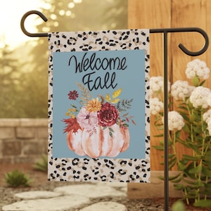 May include: A garden flag with a light blue background and the words "Welcome Fall" in black script. The flag features a watercolor pumpkin with a floral arrangement. The border has a leopard print design. The flag is displayed on a black metal stand.