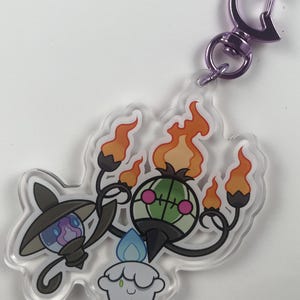 Pu&ograve; includere: Un portachiavi in acrilico trasparente con tre personaggi Pok&eacute;mon: un Chandelure verde e nero, un Litwick bianco e blu e un Lampent bianco e blu. Il portachiavi ha un moschettone in metallo viola.