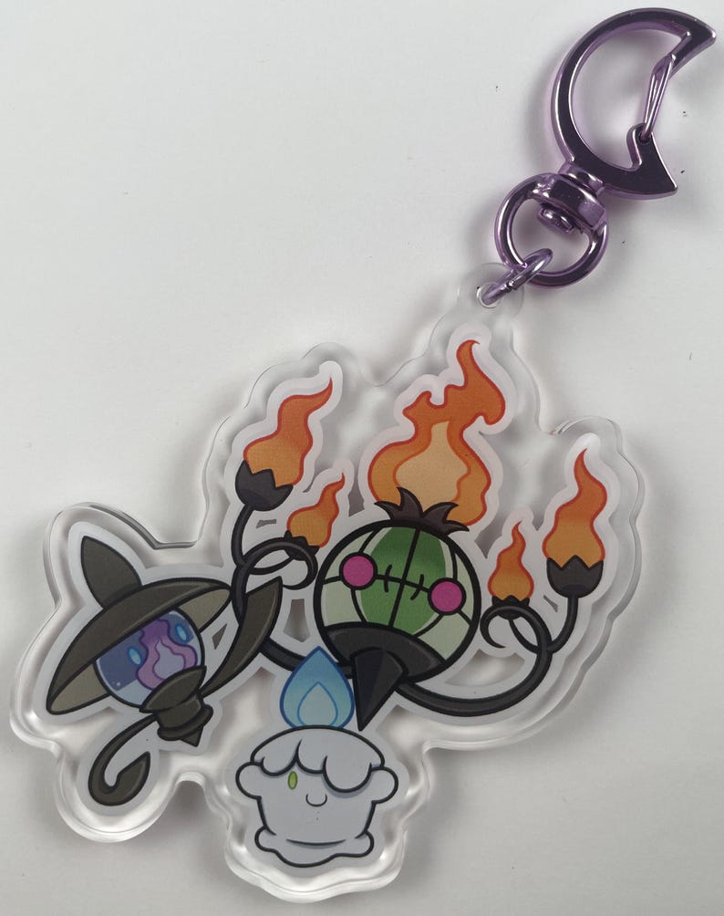 Pu&ograve; includere: Un portachiavi in acrilico trasparente con tre personaggi Pok&eacute;mon: un Chandelure verde e nero, un Litwick bianco e blu e un Lampent bianco e blu. Il portachiavi ha un moschettone in metallo viola.