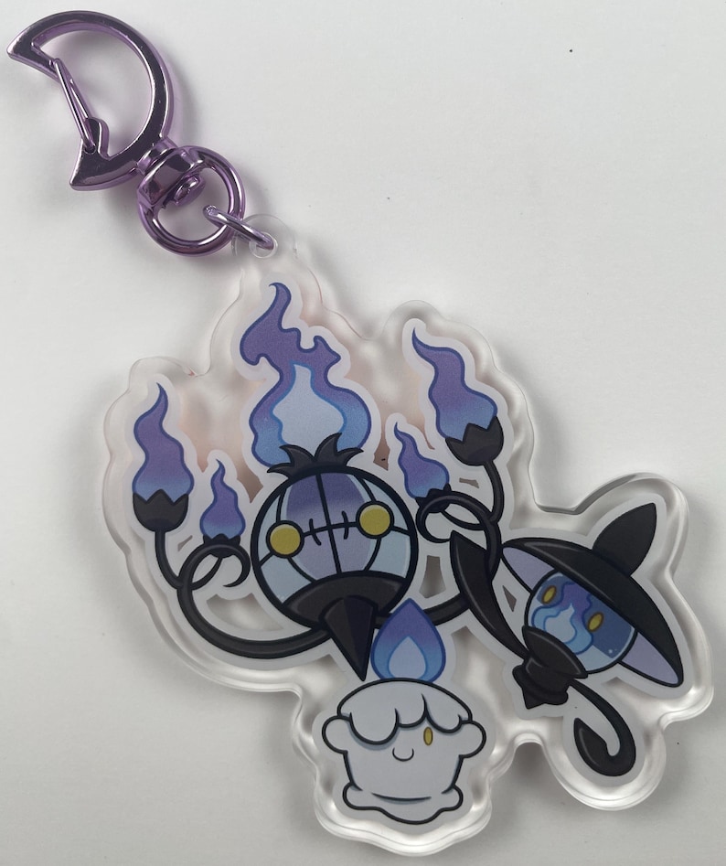 Pu&ograve; includere: Un portachiavi in acrilico viola e nero con tre personaggi Pok&eacute;mon spettrali. I personaggi sono Chandelure, Litwick e Lampent. Il portachiavi ha una chiusura metallica viola.