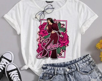 May include: White t-shirt with a graphic print of Selena Quintanilla, a singer, wearing a red outfit and holding a microphone. The graphic includes pink roses and the text "Selena" and "A Piper Valdez".