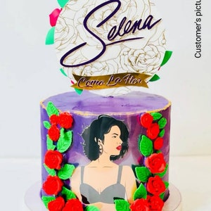 May include: Purple cake decorated with red roses and green leaves. A photo of Selena is on the cake. The cake topper says "Selena" and "Como La Flor".