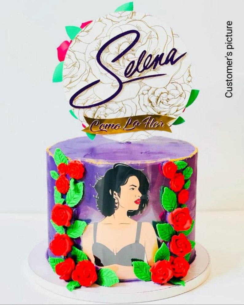 May include: Purple cake decorated with red roses and green leaves. A photo of Selena is on the cake. The cake topper says "Selena" and "Como La Flor".
