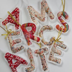 May include: Fabric letter decorations in various colours and patterns, including red floral, star, and striped designs. Each letter has a white scalloped edge and a gold ribbon for hanging. The letters spell out "ART, C, E, M, S, T, J, E, A, R".