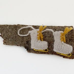 May include: Two miniature knitted ice skates in light gray with yellow laces and yellow blades are displayed on a piece of textured brown bark. The skates are connected by a loop of yarn, suggesting a decorative or ornamental purpose.