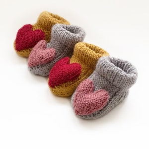 May include: Four pairs of knitted baby booties in mustard yellow, gray, and red, each featuring a heart applique. The booties are arranged diagonally on a white background, showcasing the handmade craftsmanship and soft texture of the yarn.