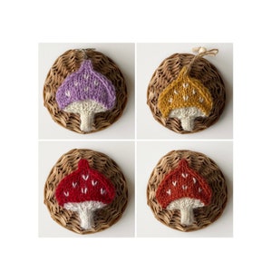 May include: Four knitted mushroom ornaments in different colors. The mushrooms are displayed on woven baskets. The mushrooms are purple, yellow, red, and orange.