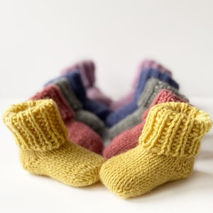 Könnte beinhalten: Eine Gruppe gestrickter Babyschuhe in verschiedenen Farben, darunter Gelb, Rosa und Lila. Die Schuhe sind in einer Reihe angeordnet, wobei die gelben Schuhe im Vordergrund stehen.