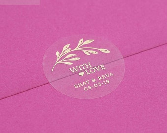 Op de afbeelding: Een roze envelop met een ronde, doorzichtige sticker met een gouden bloemenmotief en de tekst "With Love Shay & Reva 08.03.19"