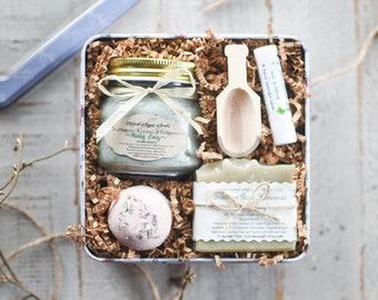 May include: A gift set in a floral tin, filled with a jar of sugar scrub, a wooden scoop, a lip balm, a bar of soap, and a bath bomb. The jar has a cream-colored label with text. The bath bomb is pink with lavender.