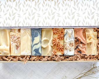 May include: A gift box filled with eight rectangular bars of artisan soap. The soaps vary in color, including white, beige, and brown, with decorative elements like swirls and dried flowers. The box has a floral patterned lid and is lined with brown paper.