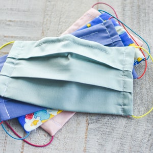 May include: A stack of four fabric face masks with elastic ear loops. The top mask is light blue, the second is purple, the third is pink with a colorful pattern, and the bottom mask is blue with a white and blue pattern.