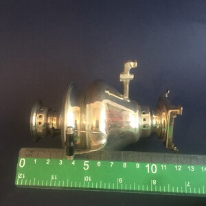 Puede incluir: Un samovar plateado con boquilla, asa y elementos decorativos. El samovar mide aproximadamente 13 cm de largo, medido con una regla verde. El fondo es azul oscuro.