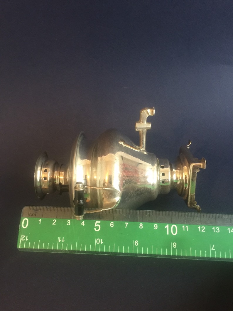 Puede incluir: Un samovar plateado con boquilla, asa y elementos decorativos. El samovar mide aproximadamente 13 cm de largo, medido con una regla verde. El fondo es azul oscuro.