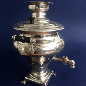 Puede incluir: Un samovar plateado con un asa y un pico negros. La tetera tiene un cuerpo redondo, una tapa y se asienta sobre una base cuadrada. Un utensilio tradicional ruso para el t&eacute;.