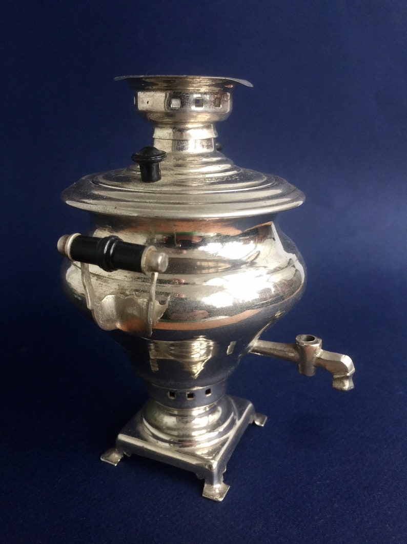 Puede incluir: Un samovar plateado con un asa y un pico negros. La tetera tiene un cuerpo redondo, una tapa y se asienta sobre una base cuadrada. Un utensilio tradicional ruso para el t&eacute;.