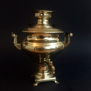 Puede incluir: Un samovar de lat&oacute;n vintage con una superficie brillante y reflectante. La tetera presenta un cuerpo redondeado, una tapa con una peque&ntilde;a chimenea y dos asas curvas. Tiene un grifo cerca de la base y se asienta sobre una base cuadrada.