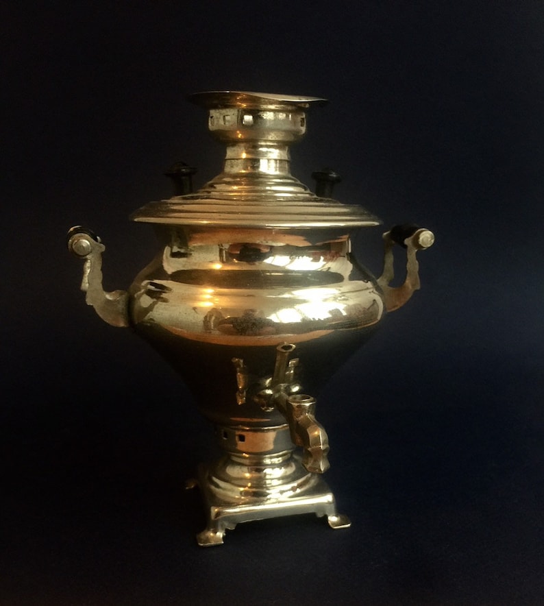 Puede incluir: Un samovar de lat&oacute;n vintage con una superficie brillante y reflectante. La tetera presenta un cuerpo redondeado, una tapa con una peque&ntilde;a chimenea y dos asas curvas. Tiene un grifo cerca de la base y se asienta sobre una base cuadrada.