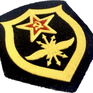 Puede incluir: Un parche de fieltro negro con un borde amarillo y una estrella roja con un martillo y una hoz en su interior. La estrella está centrada sobre una estrella estilizada amarilla con alas.